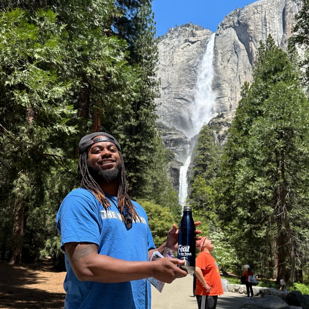man smiling holding a water bottle with waterfall and trees in background
