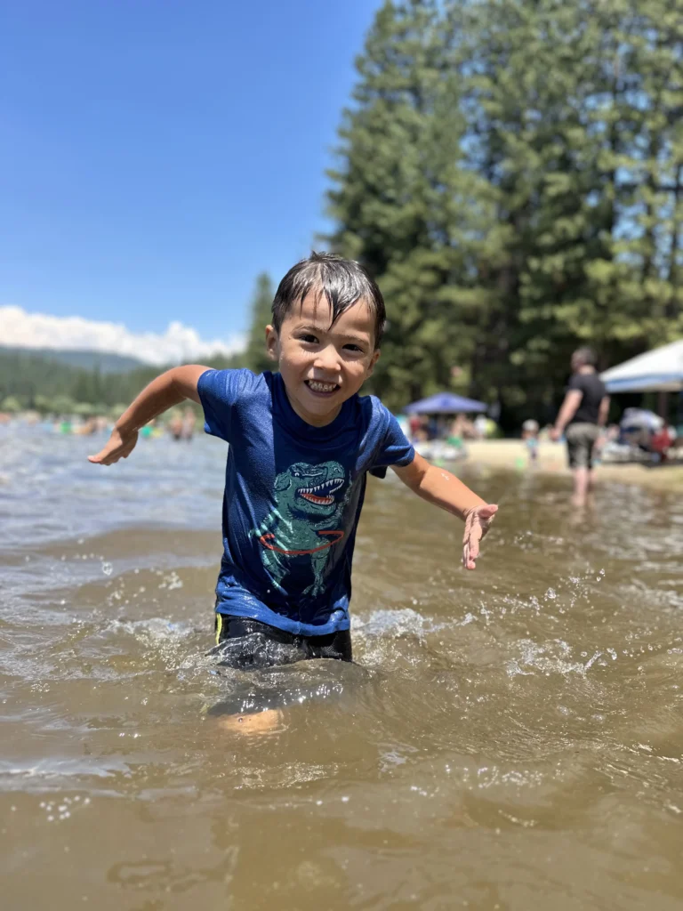 child smiling and playing in the lake water with trees and popup shades on beachfront in background