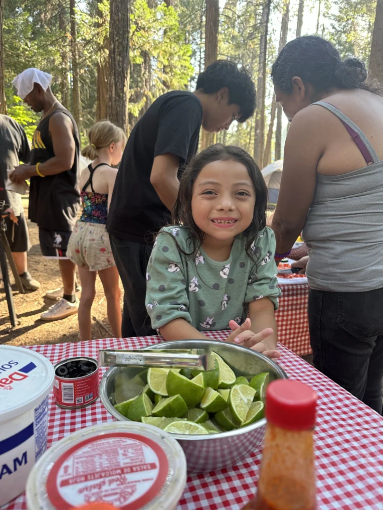 girl smiling at camera with bowl of limes in foreground on table and people preparing food in background with trees behind