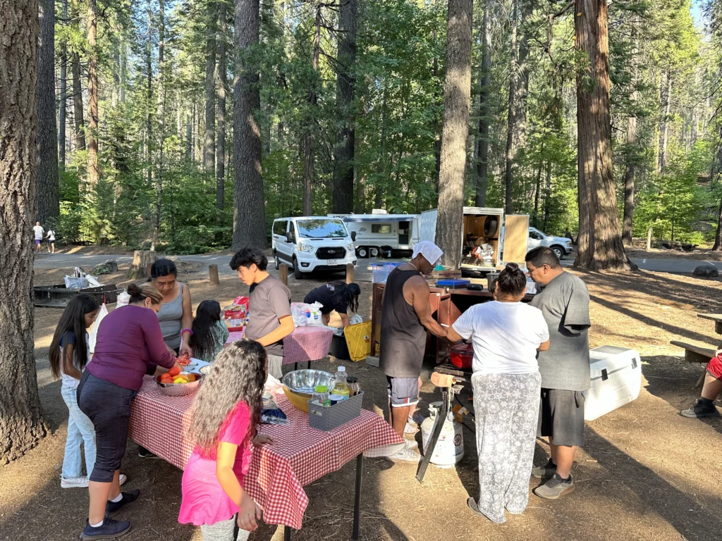 group of people preparing food around tables at a campsite with vehicles in background around the trees