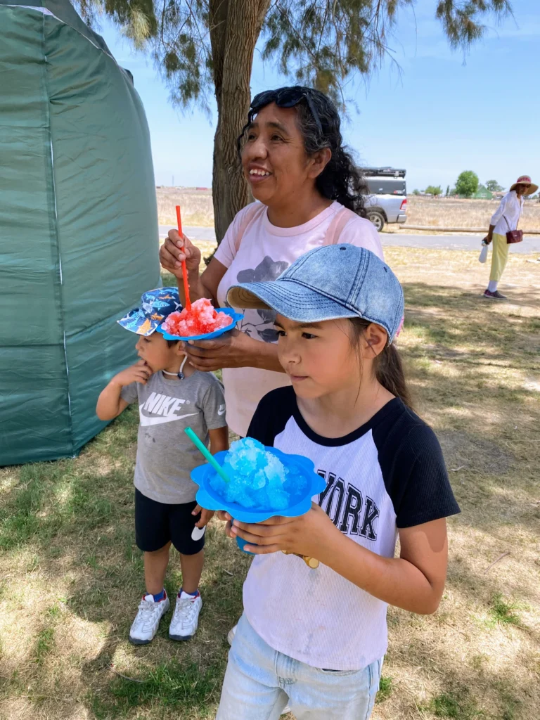 woman and two children eating snow cones beneath shady tree