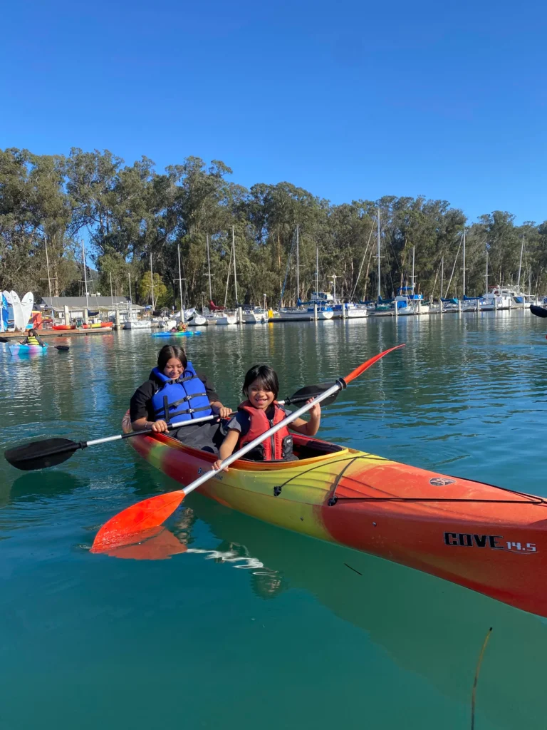two kids smiling and paddling in calm water with boats docked in the background