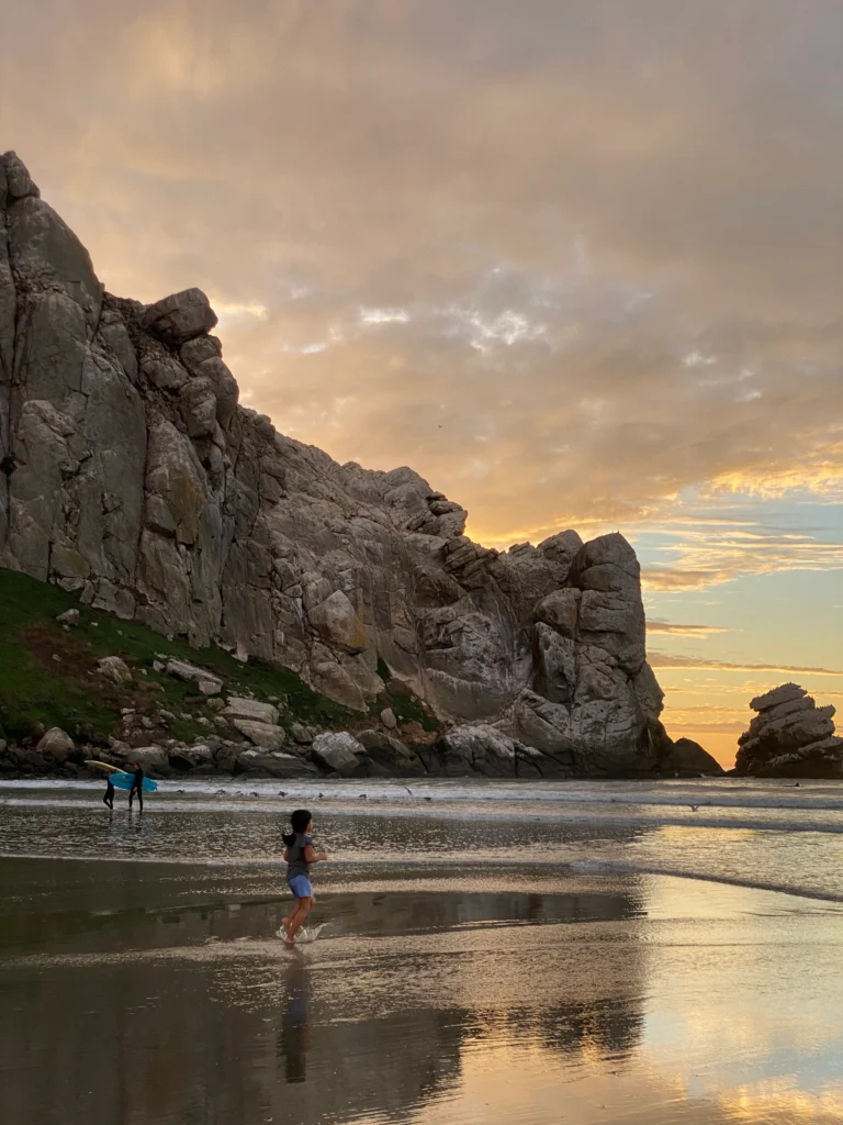 kid running in the water at the beach with sunset behind rocks jutting into the ocean