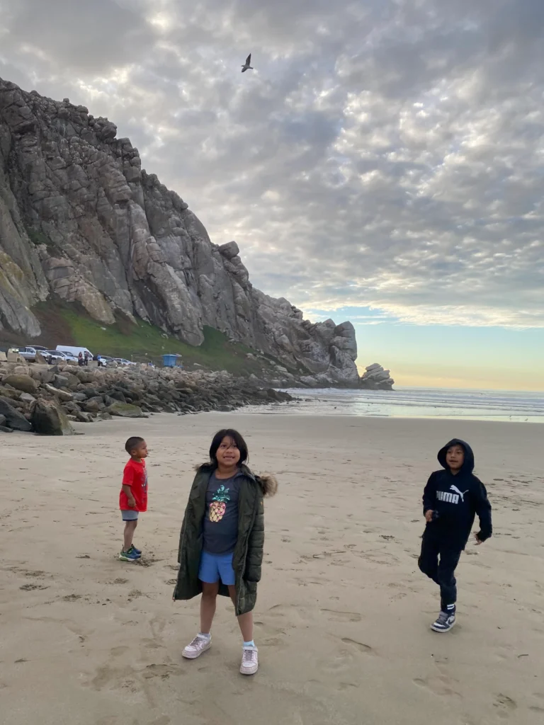 three kids standing and smiling on beach with rock outcropping going into water in background