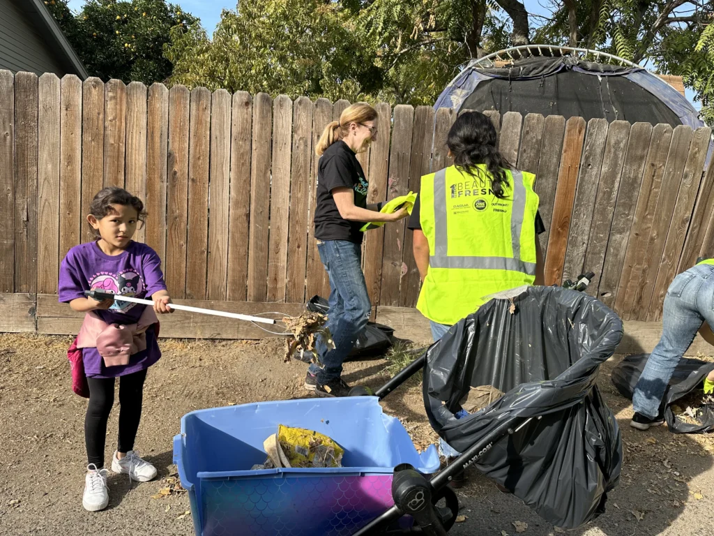 two adults and a kid cleaning up an alleyway along a fence line