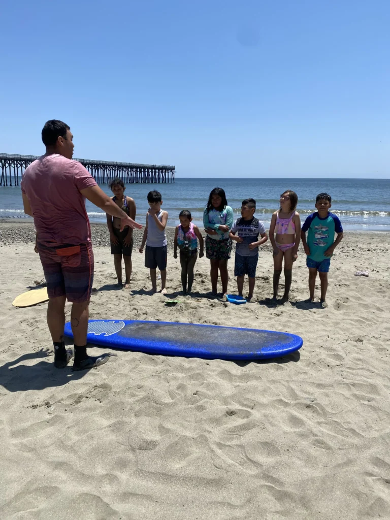 man standing in front of children with surfboard on sand