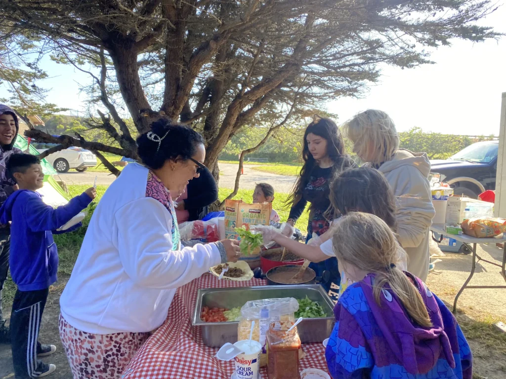 people gathering around food on picnic table under a tree on a sunny day